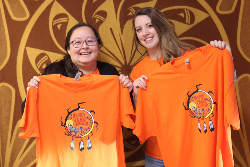 Two students proudly displaying vibrant orange shirts featuring a custom design created by nehiyaw artist and NWP alumni, Diane Meneen