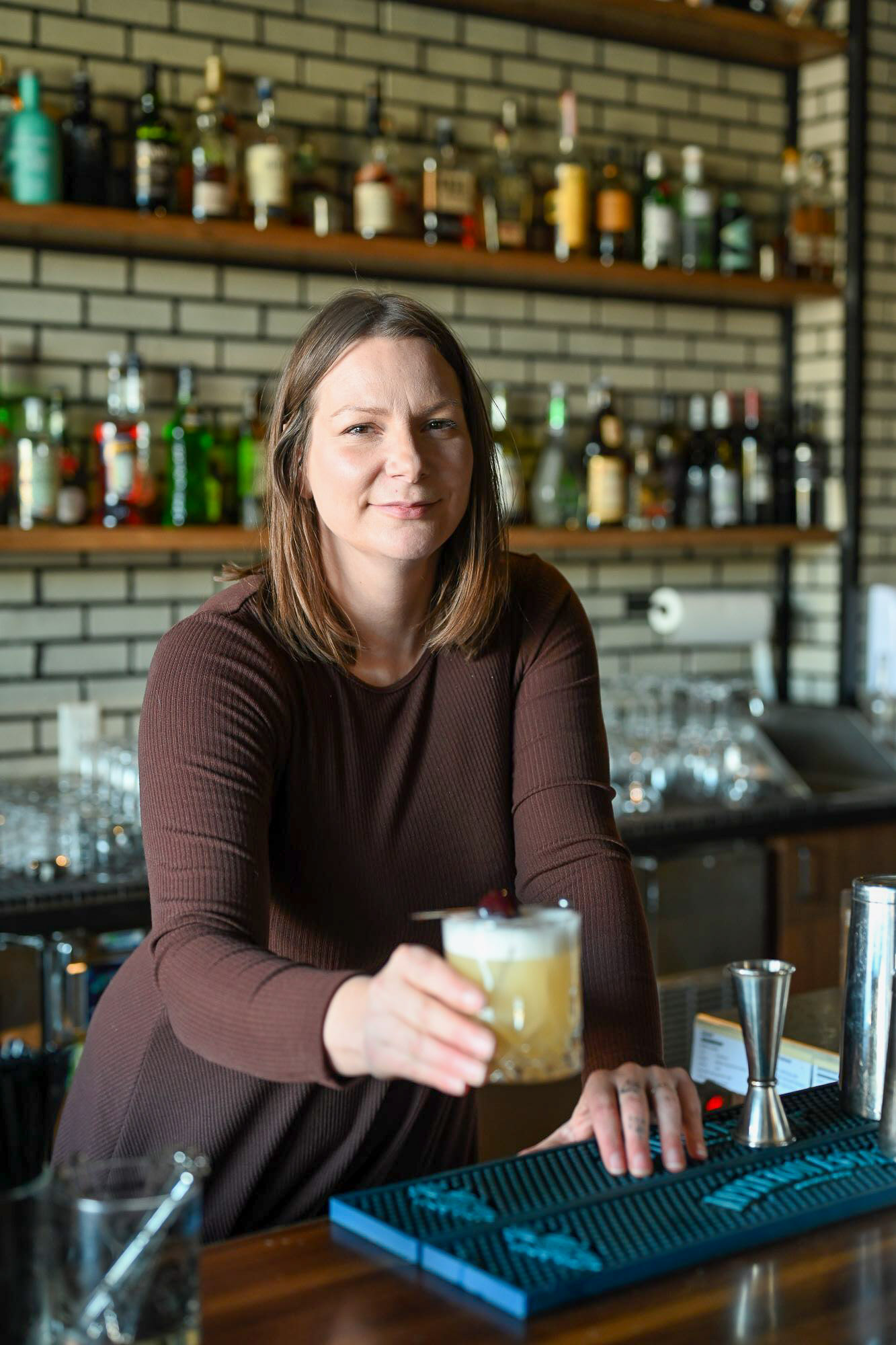Jessica Schoepp, 92 Beverage Co owner and NWP Alumna holding a delicious cocktail behind the bar with an engaging collection of liquor in the background