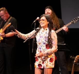 NWP Alumni Melody McArthur on stage holding up a microphone with her band in the background.