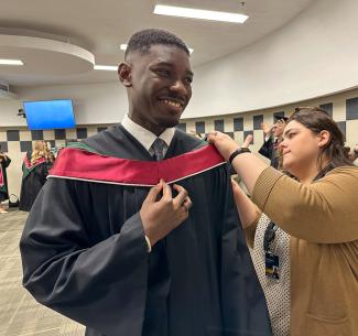2025 Valedictorian Clinton Owusu smiles while receiving assistance gowning up before the Convocation ceremony