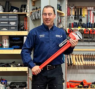 NWP plumbing and gasfitter instructor, Richard Bell holding a large wrench, posing in front of a tool cupboard.