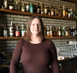 Jessica Schoepp, 92 Beverage Co owner and NWP Alumna standing behind the bar with an engaging collection of liquor in the background