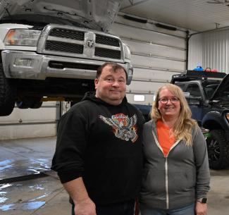 Danielle and Stacey Commandeur stand together inside their automotive and heavy-duty repair shop, with two pickup trucks behind them in the service area.