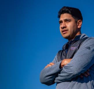 Mens soccer head coach, David Rojas observing the team play with a bright, clear blue sky in the background
