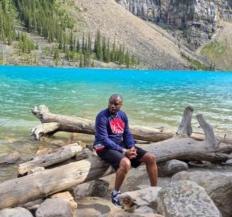 Phil Mvondo seated on a large tree trunk near an aquamarine-colored lake, surrounded by the stunning landscape of the Canadian Rockies.