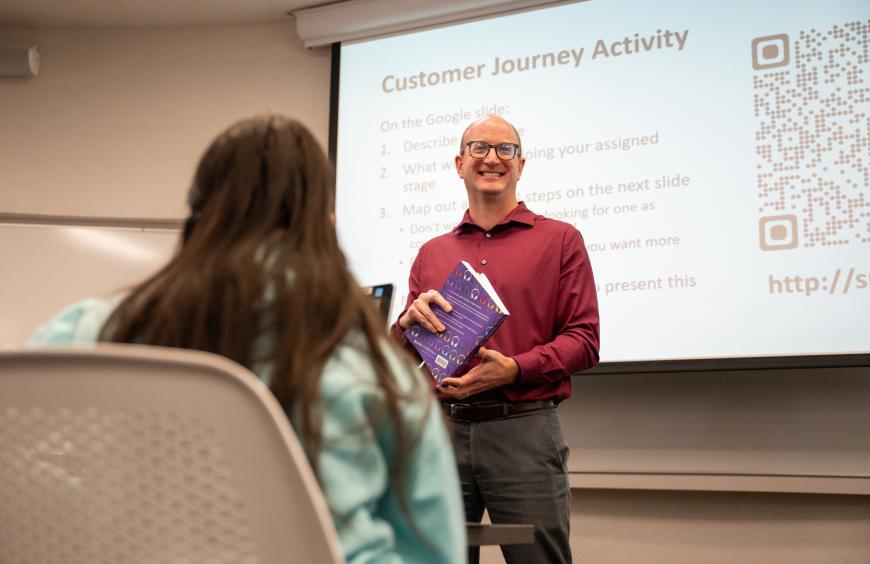A smiling instructor standing at the front of a class with a book in his hand