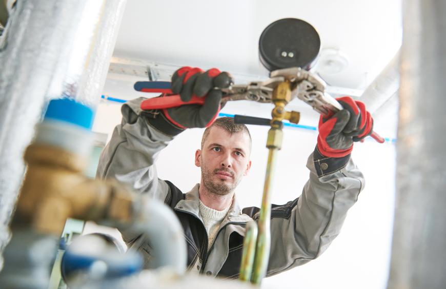 Tradesperson in a gray jacket and gloves gripping a pipe with a large wrench.