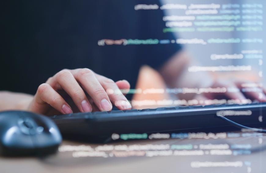 A view of hands typing on a keyboard with computer code displayed on a monitor.