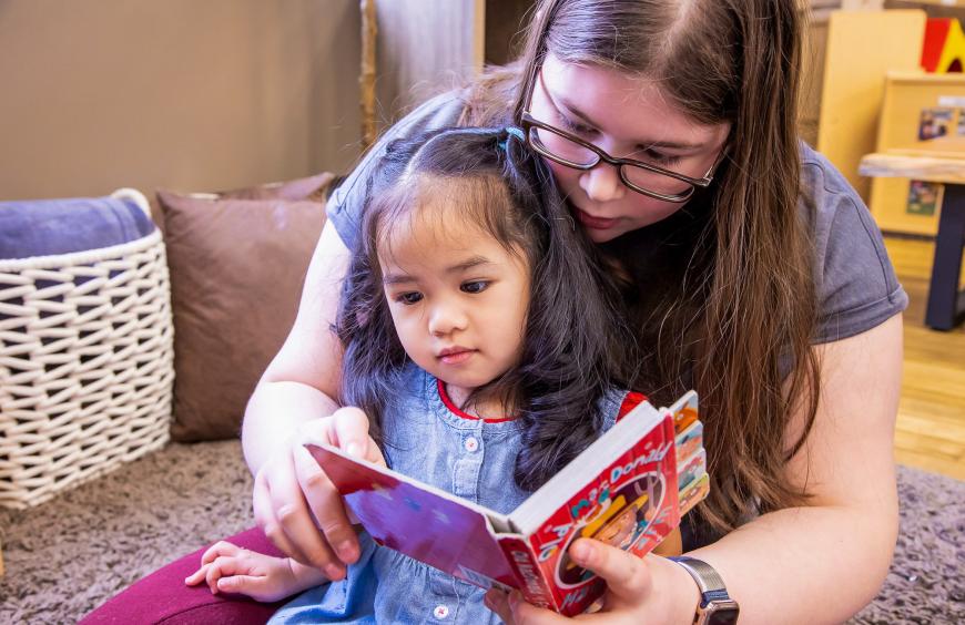 An early childhood educator sitting with an infant reading a book in a preschool