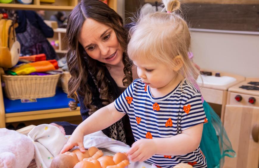 Early childhood educator encouraging a toddler who is playing with a pair of dolls