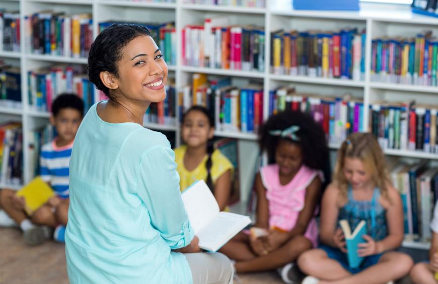 A happy teacher sitting in front of a group of young children in a classroom with bookshelves in the background