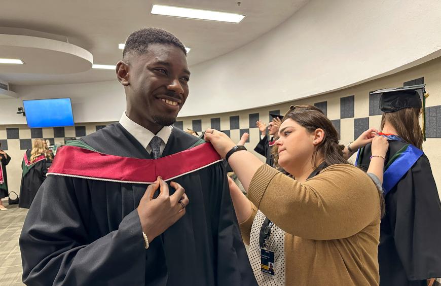 2025 Valedictorian Clinton Owusu smiles while receiving assistance gowning up before the Convocation ceremony