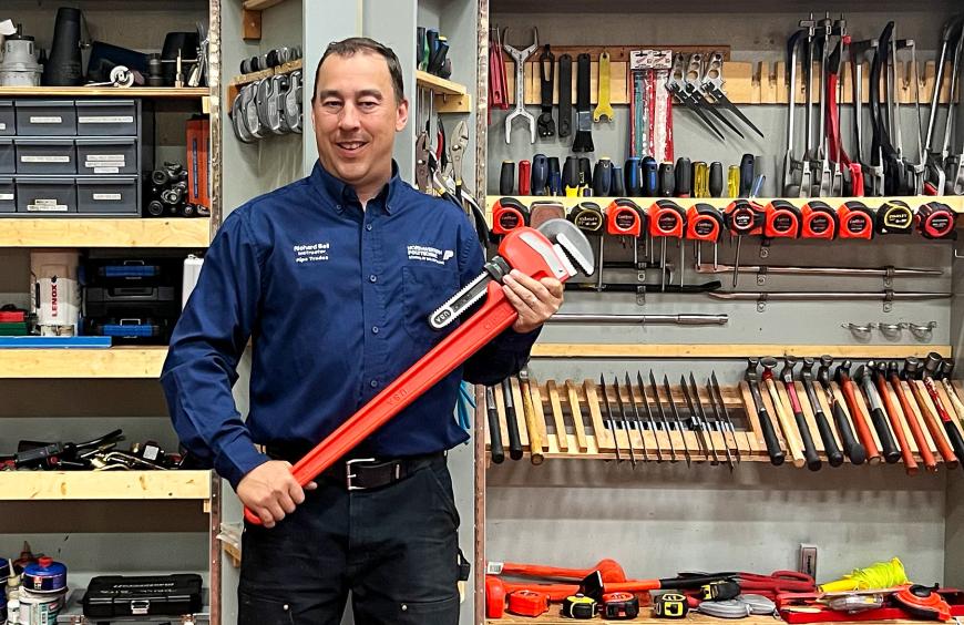NWP plumbing and gasfitter instructor, Richard Bell holding a large wrench, posing in front of a tool cupboard.