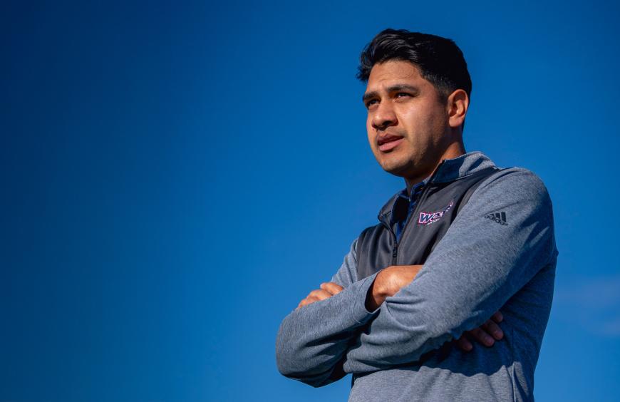 Mens soccer head coach, David Rojas observing the team play with a bright, clear blue sky in the background