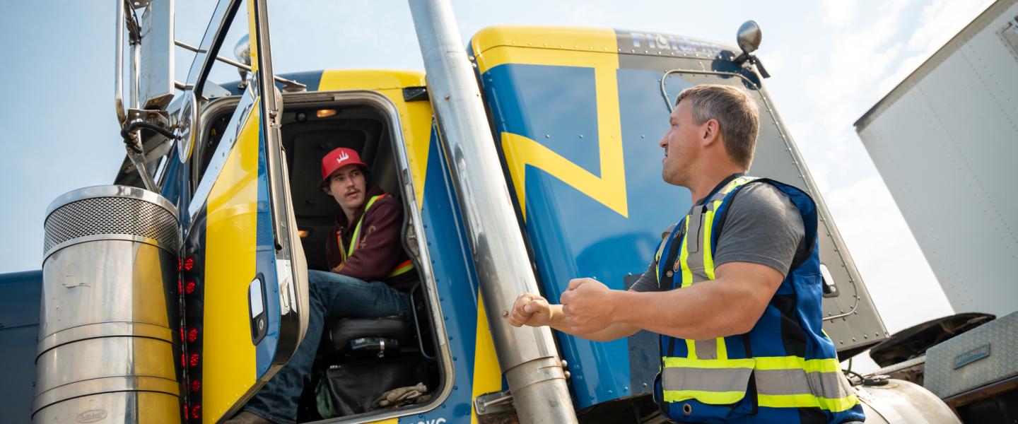 Student sitting in the cab of a semi-truck receiving instruction from a driving instructor
