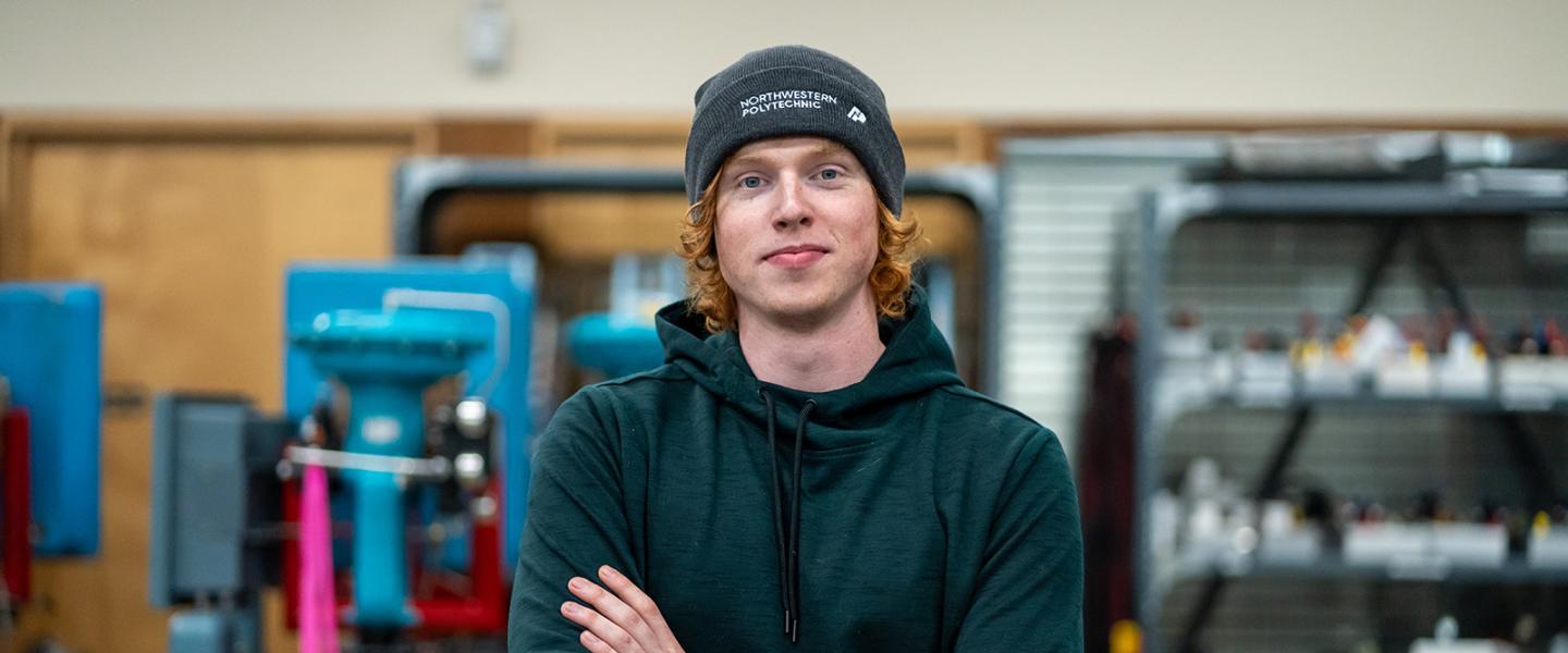 Student wearing a toque posing with arms folded in a workshop with machinery and tools in the background