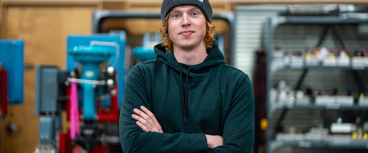 Student wearing a toque posing with arms folded in a workshop with machinery and tools in the background