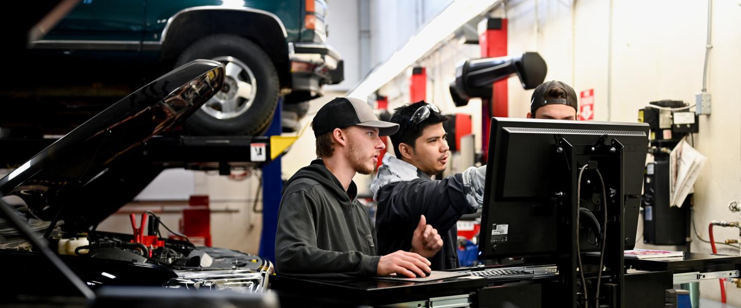 3 men collaborating around a computer in an auto repair shop surrounded by tools and and a lifted vehicle