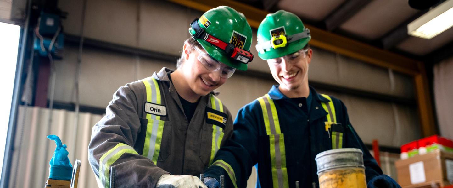 Two tradesmen wearing coveralls and hard hats share a laugh while working on equipment