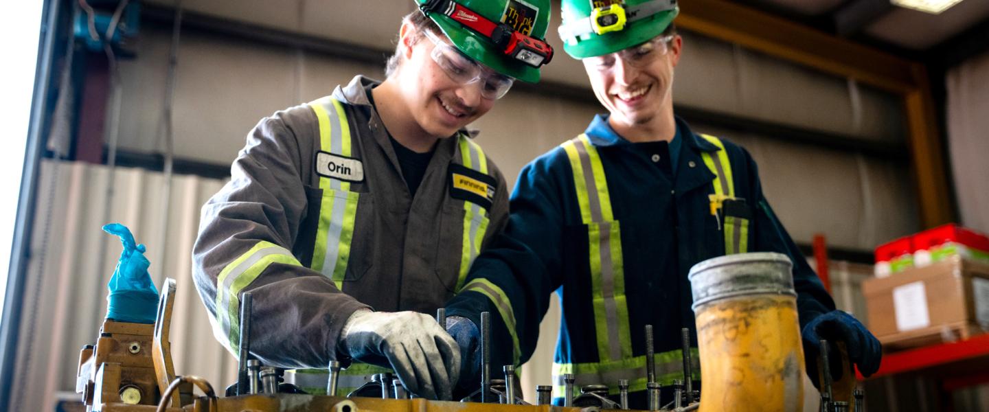 Two tradesmen wearing coveralls and hard hats share a laugh while working on equipment