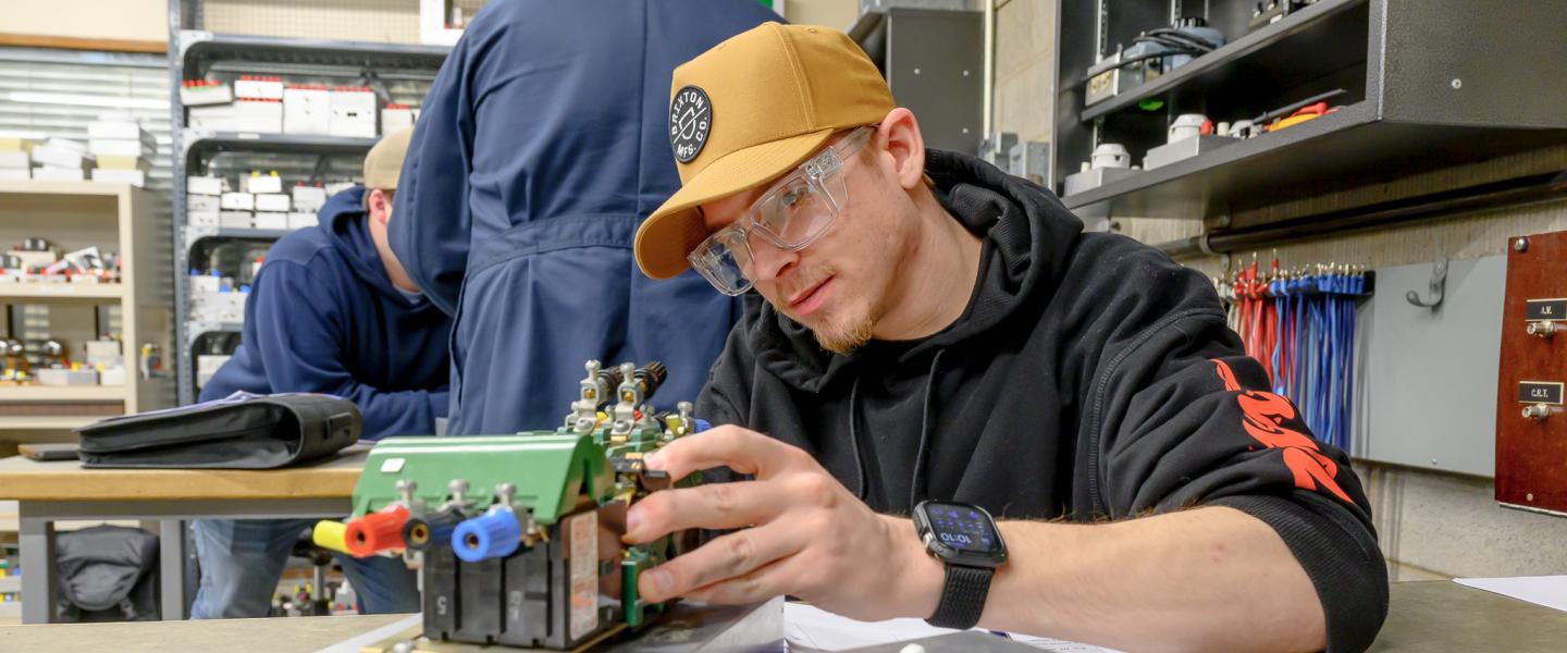 An electrician student wearing safety googles closely observing a piece of electrical equipment in the electronics lab at NWP