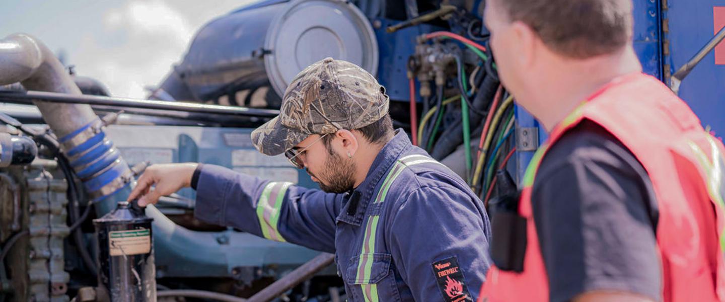 A driving instructor observes a student checking the engine of a semi-truck.