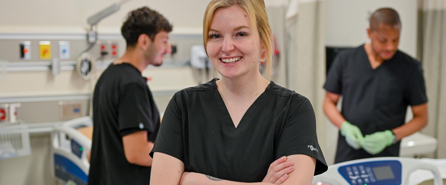 A happy practical nursing student in the simulation lab, posing with arm folded as fellow students work in the background.