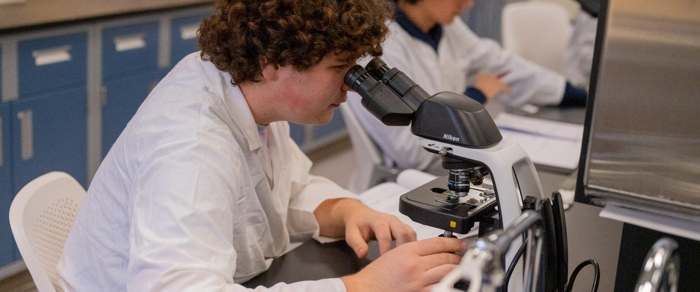 A student wearing a labcoat in the biology lab looking through a microscope
