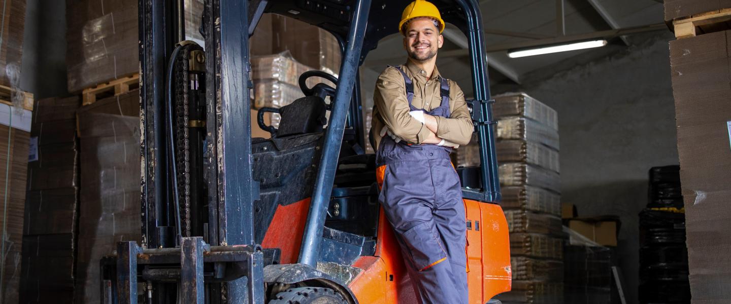 Warehouse employee wearing safety gear leaning against a forklift in a warehouse stacked high with moving boxes