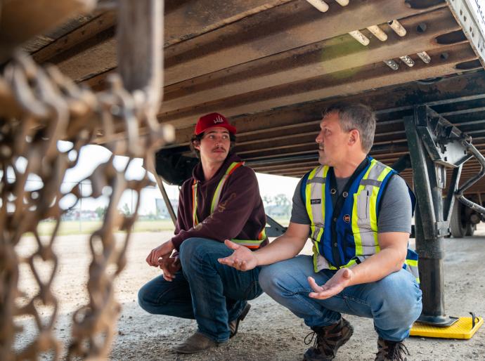 Student receiving instruction from a driving instructor underneath a semi truck