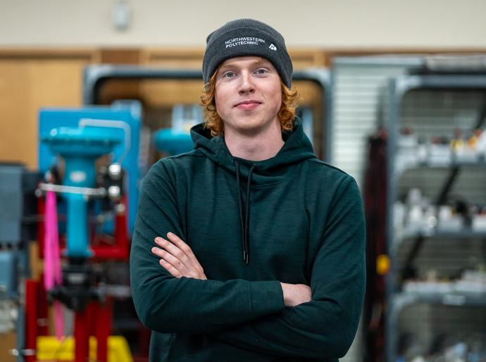 Student wearing a toque posing with arms folded in a workshop with machinery and tools in the background