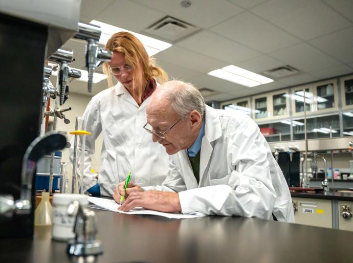 A student and organic chemistry instructor in lab coats collaborate on paperwork at a desk, focused on their research and findings