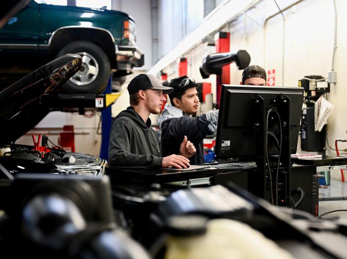 3 men collaborating around a computer in an auto repair shop surrounded by tools and and a lifted vehicle