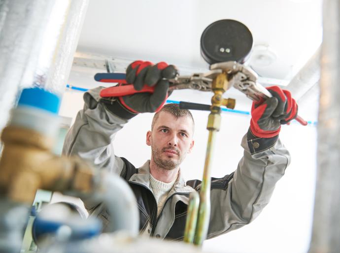 Tradesperson in a gray jacket and gloves gripping a pipe with a large wrench.