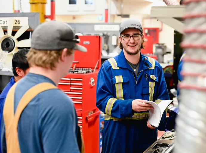 Two men in blue overalls chatting in a workshop with tools in the background.