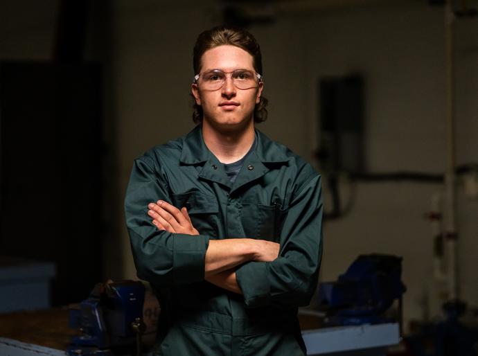 Tradesperson wearing protective eye wear and coveralls, standing in a workshop with arms folded.