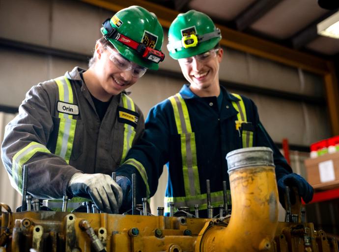 Two tradesmen wearing coveralls and hard hats share a laugh while working on equipment