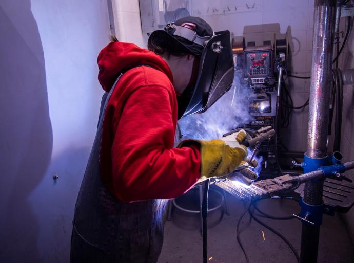 A person equipped with a welding helmet and gloves is engaged in welding, surrounded by bright sparks and metal.