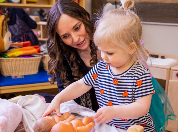 Early childhood educator encouraging a toddler who is playing with a pair of dolls