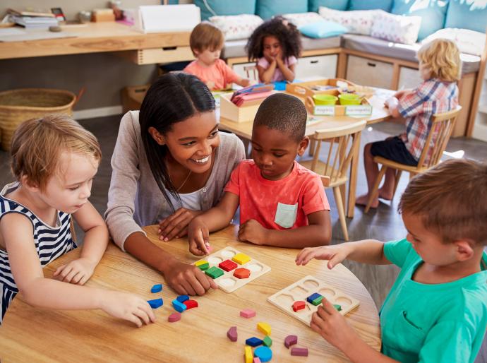 An happy early childhood educator in a classroom engaging with young children sitting around a crafting table playing a game.
