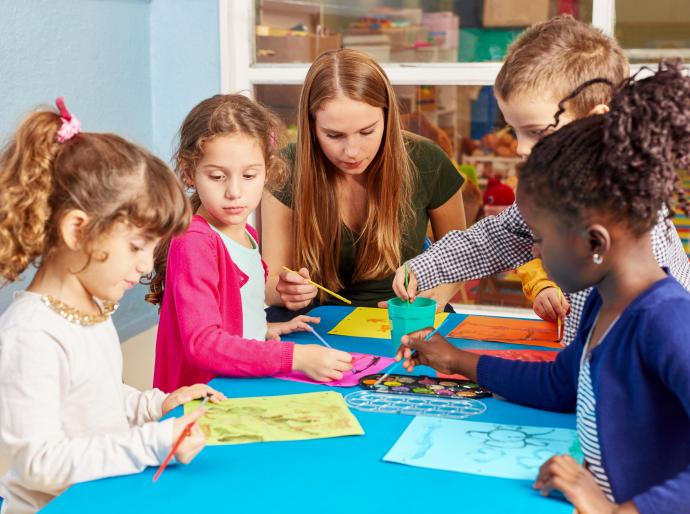 A teacher interacting with a group of children who are drawing pictures on a crafting table