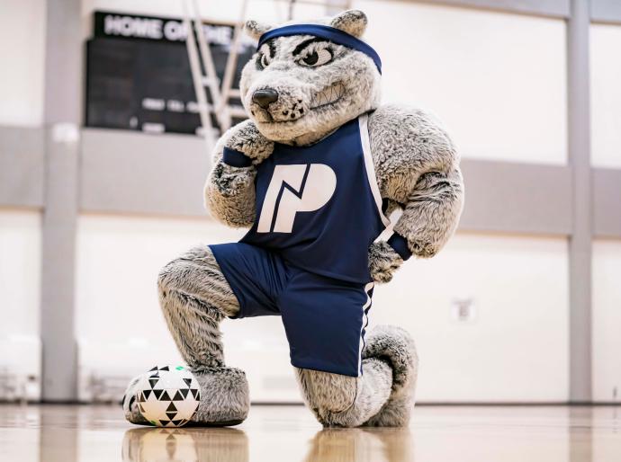 Farley the mascot kneeling in the gym with a soccer ball at his feet