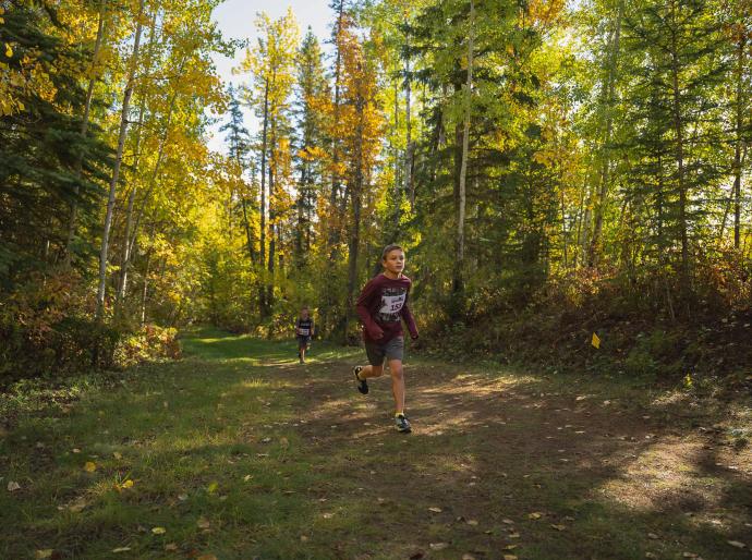 A young athlete running along a trail with trees in full fall colours in the background