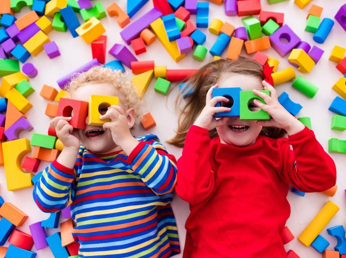 Two happy children laying down and holding building blocks up to their eyes and surrounded by lots of colourful blocks