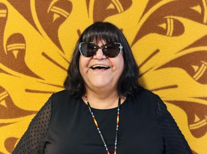 Portrait of Elder Theresa happily smiling in front of the Indigenous blanket that hangs outside the on-campus friendship centre
