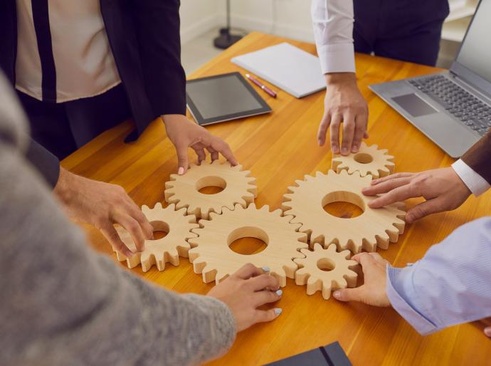 Company workers standing around a table joining cogwheels as metaphor for teamwork, business system and working solutions