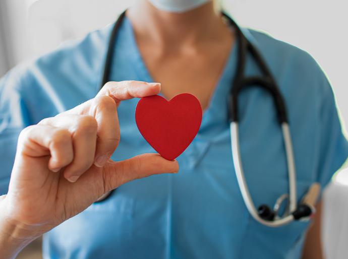 A nurse wearing blue scrubs with a stethoscope around her neck holding up a red heart between her thumb and index finger