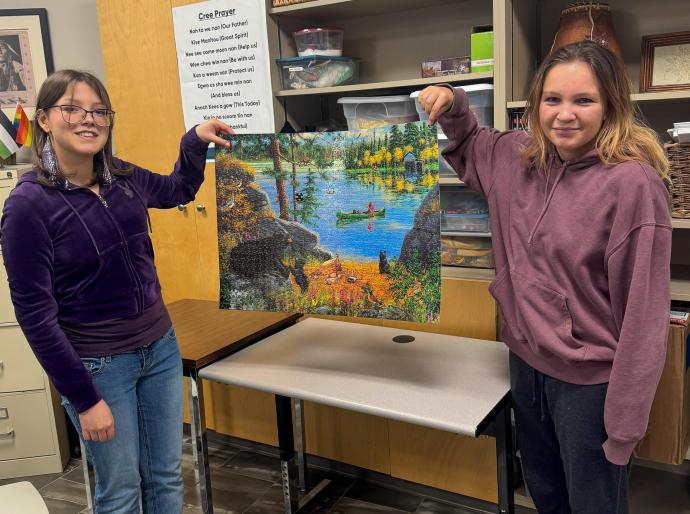 Two students holding up a completed  jigsaw puzzle of a person in a canoe on a beautiful blue lake