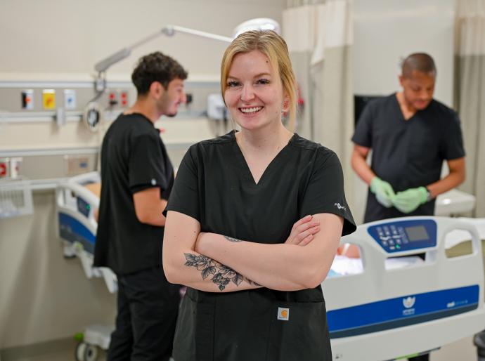 A happy practical nursing student in the simulation lab, posing with arm folded as fellow students work in the background.