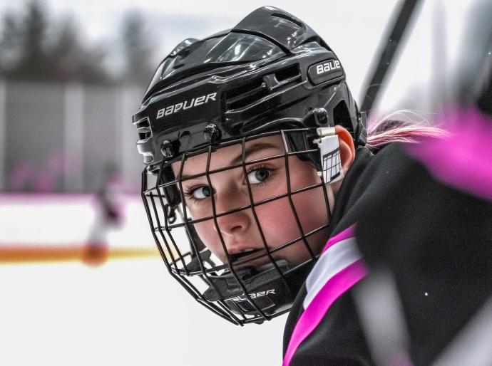 A female hockey player wearing a helmet looking directly at the camera from the team bench